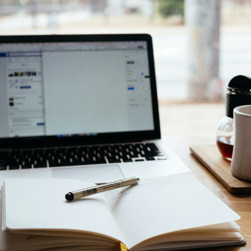 Photo of a pen on a notebook with a laptop behind it next to a cup of coffee.